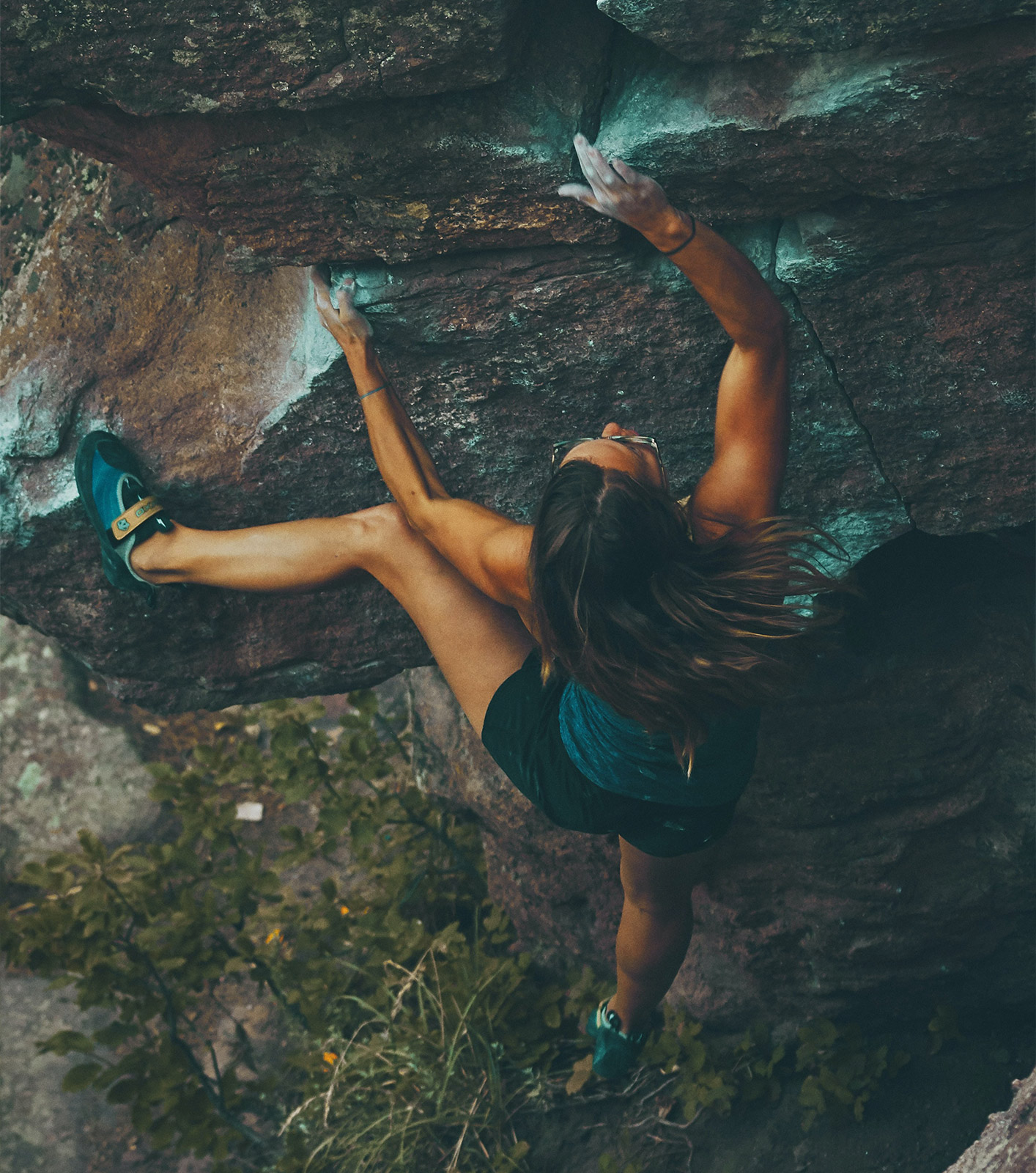 Donna, abbronzata, con capelli lunghi scuri, occhiali, scarpe da arrampicata mentre fa bouldering su roccia.