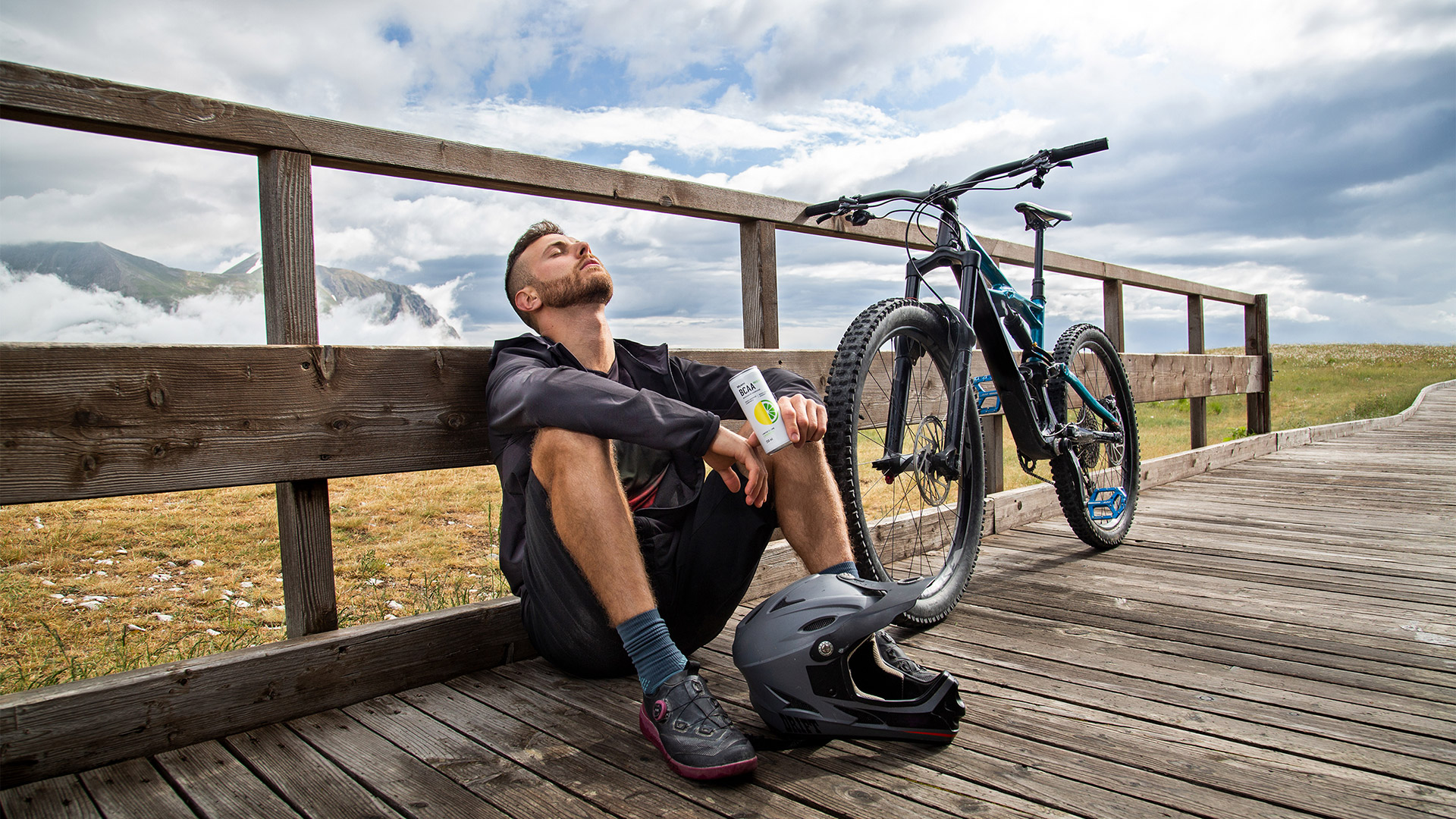 Uomo seduto vicino a una mountainbike con una bevanda BCAA in lattina in mano ed un elmo da mountainbike di fronte ai suoi piedi