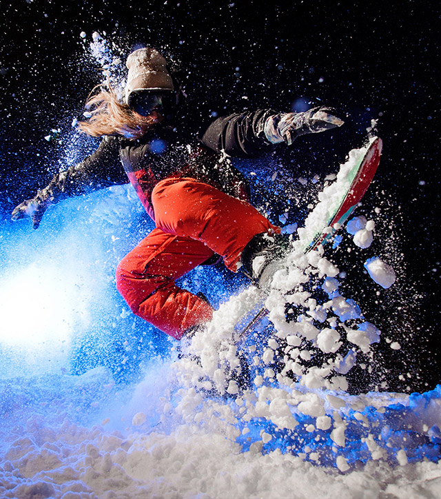 Una persona con i capelli lunghi che indossa abiti da sport invernali, cappello, guanti e maschera da sci salta con lo snowboard, sollevando la neve.